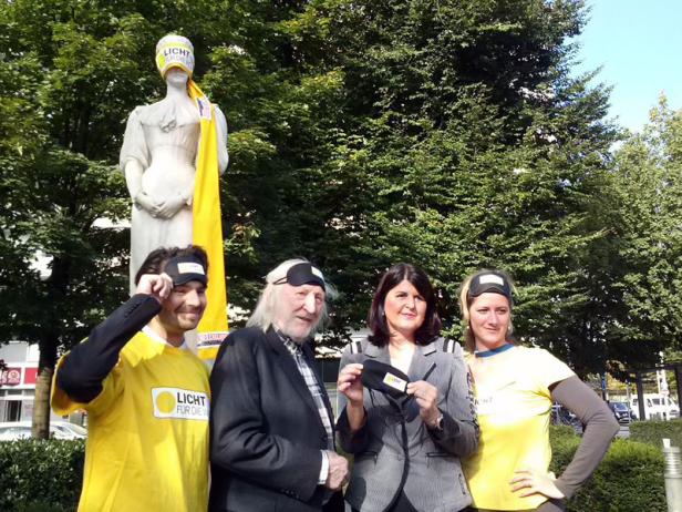 Eine Gruppe von Menschen steht vor einer Statue mit Augenbinde und gelbem Tuch.