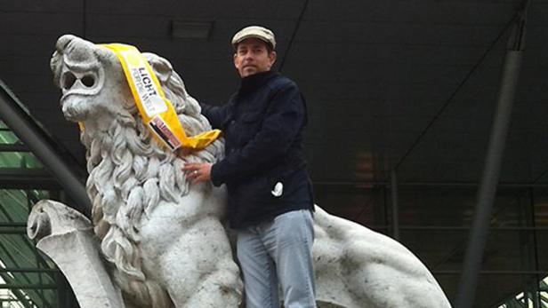 Ein Mann posiert neben einer Löwenstatue mit einer gelben Schärpe mit der Aufschrift „Licht für die Welt“.