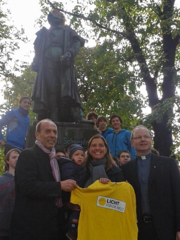 Eine Gruppe von Menschen steht vor einem Denkmal und hält ein gelbes T-Shirt mit der Aufschrift „Licht für die Welt“.