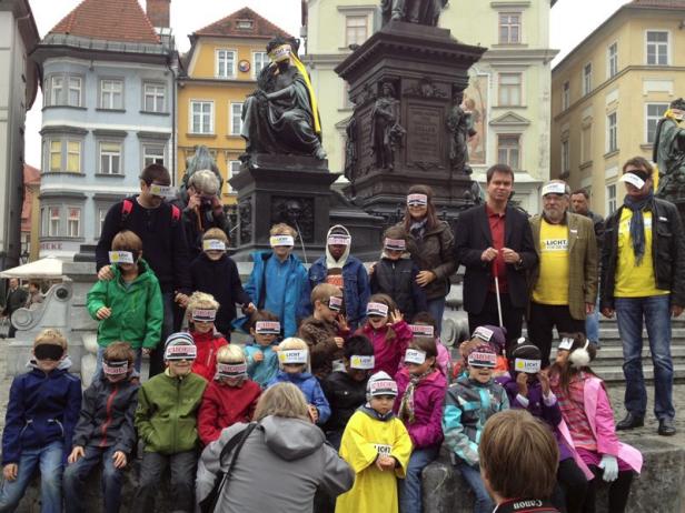 Eine Gruppe von Kindern und Erwachsenen mit Augenbinden vor dem Denkmal Erzherzog Johann in Graz.