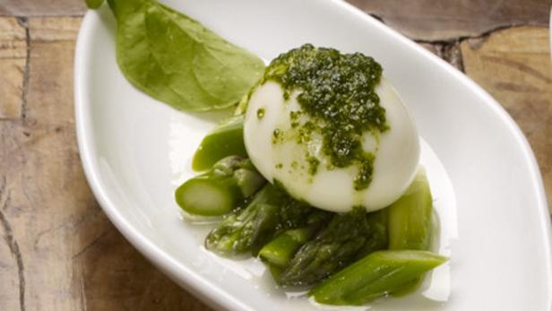 Ein pochiertes Ei mit Pesto auf Spargel und einem Blatt Basilikum auf einem weißen Teller.