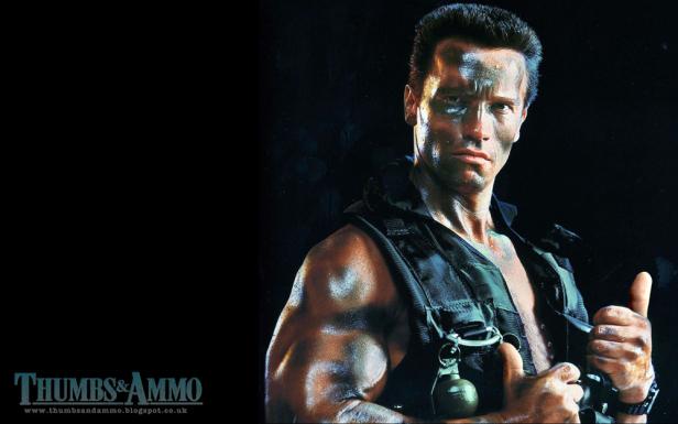 Arnold Schwarzenegger als muskulöser Soldat mit Kriegsbemalung und einer Granate an seiner Weste gibt einen Daumen nach oben.