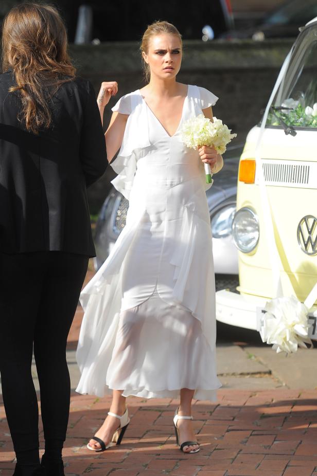 Cara Delevingne in einem weißen Kleid mit Blumenstrauß vor einem VW-Bus.