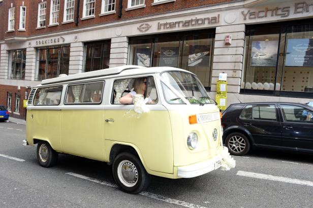 Ein gelber VW-Bus, geschmückt mit weißen Blumen, fährt durch eine Stadt.