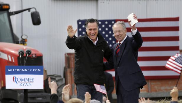 Mitt Romney und ein weiterer Mann winken vor einer amerikanischen Flagge und einem Traktor.