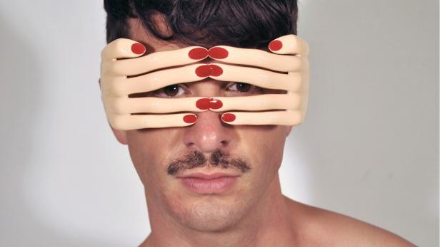 Ein Mann trägt eine Maske, die aus mehreren Händen mit rot lackierten Nägeln besteht.