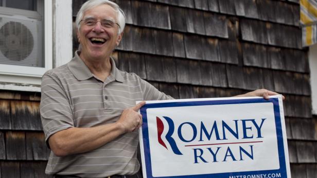 Ein lachender Mann mit Brille zeigt auf ein Wahlplakat für Romney und Ryan.