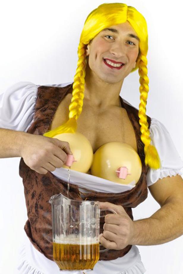 Ein Mann im Dirndl mit gelber Perücke gießt Bier aus einer Attrappe-Brust in einen Krug.