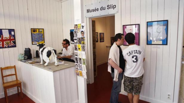 Besucher betrachten Kunstwerke im „Musée du Slip“, während ein Mann am Empfang sitzt.