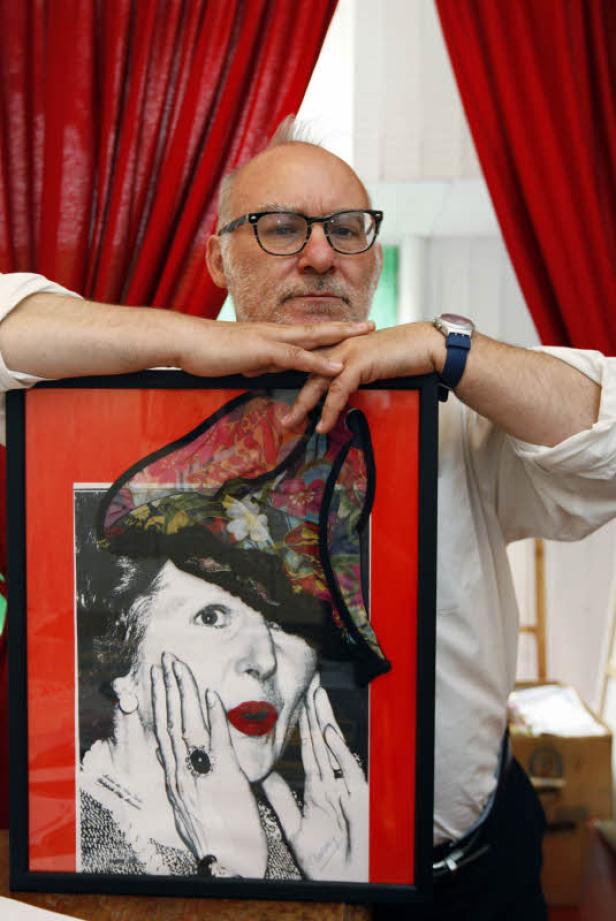 Ein Mann mit Brille lehnt an einem gerahmten Porträt einer Frau mit Hut.