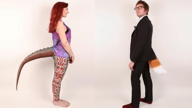 Eine Frau mit einem Dinosaurierschwanz und ein Mann mit einem Fuchsschwanz stehen vor einer weißen Wand.