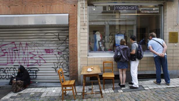 Eine Frau sitzt vor einem mit Graffiti besprühten Rollladen, während Passanten ein Schaufenster von „mana.art“ betrachten.