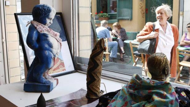 Ein Schaufenster mit einer blau-weißen Statue eines urinierenden Jungen und anderen Objekten, die Passanten reflektieren.