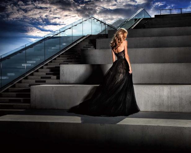 Eine Frau in einem schwarzen Kleid steht auf einer Treppe vor einem dramatischen Himmel.