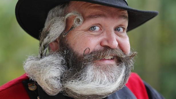 Ein Mann mit Hut und kunstvoll gezwirbeltem Bart lächelt in die Kamera.