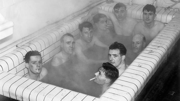 Eine Gruppe von Männern badet in einer mit Fliesen ausgelegten Wanne, einige rauchen Zigaretten.