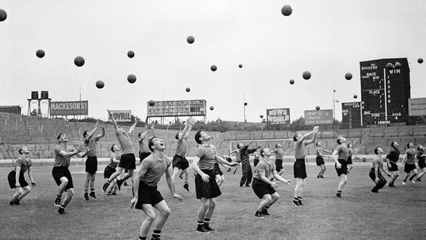 Eine Gruppe von Männern fängt Bälle auf einem Sportplatz in Schwarzweißoptik.