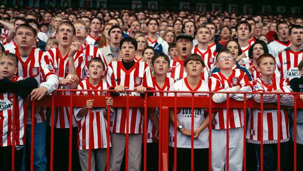 Eine große Gruppe Fußballfans, viele davon in rot-weiß gestreiften Trikots, schauen aufmerksam zu.
