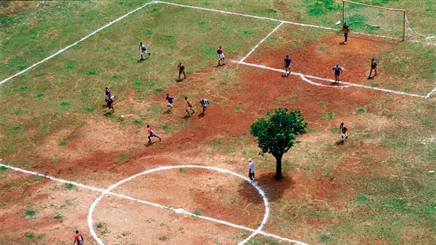 Luftaufnahme eines Fußballspiels auf einem staubigen Platz mit einem Baum in der Mitte.