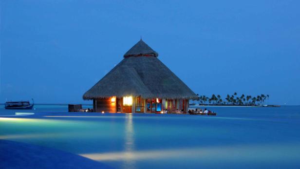 Ein strohgedecktes Gebäude auf dem Wasser, beleuchtet in der Dämmerung.