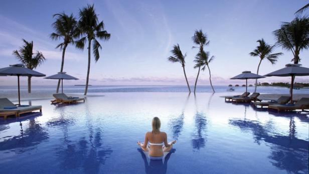 Eine Frau meditiert in einem Pool mit Blick auf Palmen und das Meer.