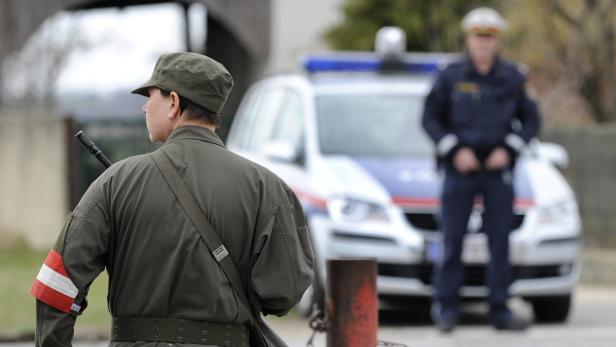 Ein Soldat mit einer Armbinde in den österreichischen Nationalfarben steht vor einem Polizeiauto.