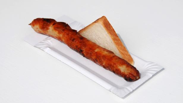 Eine Bratwurst liegt zusammen mit einer Scheibe Toastbrot auf einem Pappteller.