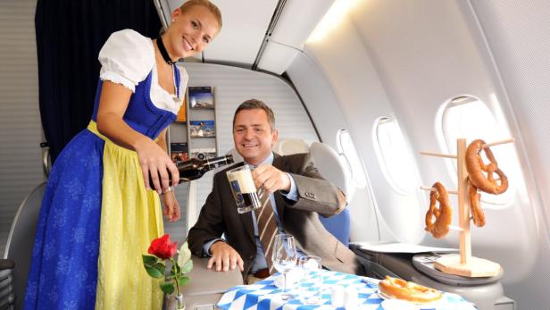 Eine Frau im Dirndl serviert einem Mann im Flugzeug Bier und Brezeln.