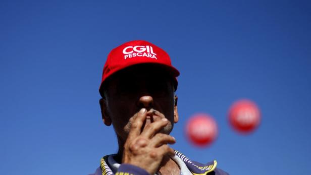 Ein Mann mit einer roten CGIL-Kappe raucht eine Zigarette vor blauem Himmel.