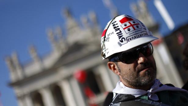 Ein Mann mit Sonnenbrille und Bauhelm mit sardischen Flaggen und Aufschriften.