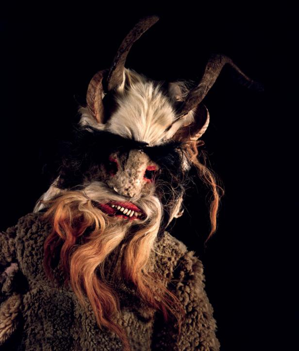 Eine Krampus-Maske mit Hörnern, Fell und roten Zähnen vor schwarzem Hintergrund.