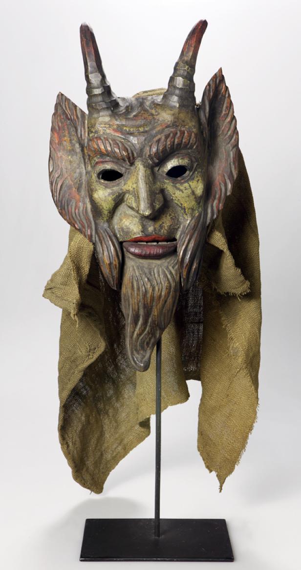 Eine geschnitzte Krampusmaske mit Hörnern und einem groben Stofftuch auf einem Ständer.