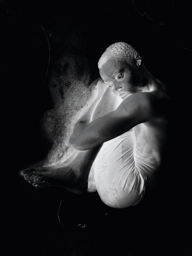 Ein Mann mit hellem Haar sitzt im Dunkeln und umarmt seine Knie.