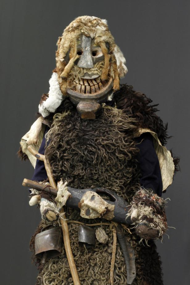 Eine traditionelle, fellbedeckte Maske mit grinsendem Gesicht und Kuhglocken an einem Kostüm.