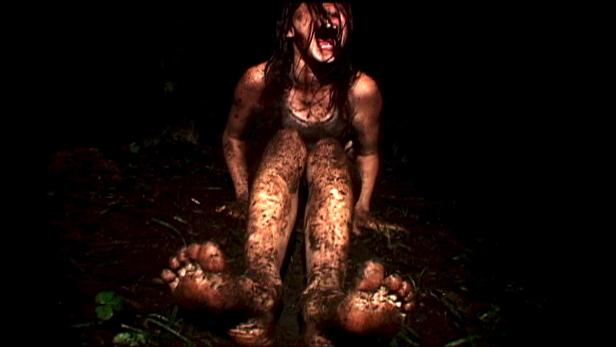 Eine schreiende, schmutzbedeckte Frau sitzt im Dunkeln auf dem Boden.