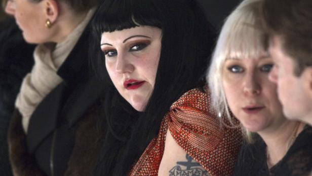 Eine Frau mit dunklen Haaren und Pony, rotem Lippenstift und einem Tattoo am Oberarm sitzt im Publikum.