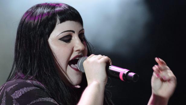 Eine Sängerin mit dunklen Haaren und auffälligem Make-up singt in ein Mikrofon.