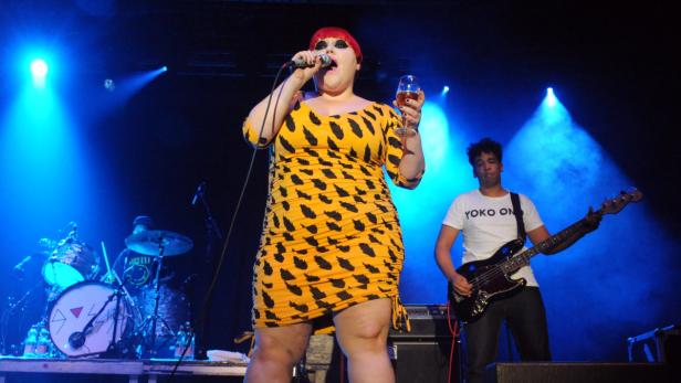 Eine Sängerin in einem gelb-schwarzen Kleid singt mit einem Mikrofon und hält ein Glas Wein auf einer Bühne.