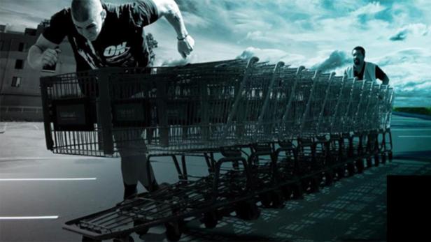 Ein Mann schiebt einen langen Zug von Einkaufswagen vor sich her, während ein anderer Mann hinten steht.