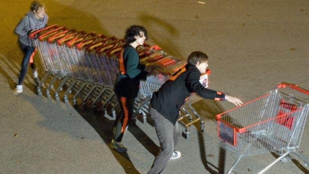 Drei Personen schieben eine lange Reihe von Einkaufswagen auf einem Parkplatz.