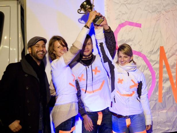 Eine Gruppe von vier Personen feiert mit einem goldenen Pokal vor einem dekorierten Hintergrund.