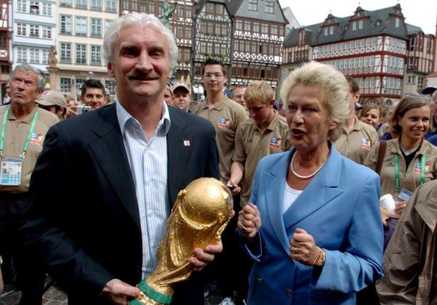 Ein Mann hält den FIFA-Pokal in der Hand, während eine Frau daneben gestikuliert; im Hintergrund Fachwerkhäuser.