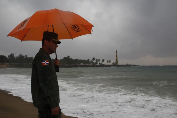 Ein Mann in Uniform steht mit einem Regenschirm am Strand; im Hintergrund ein Leuchtturm.
