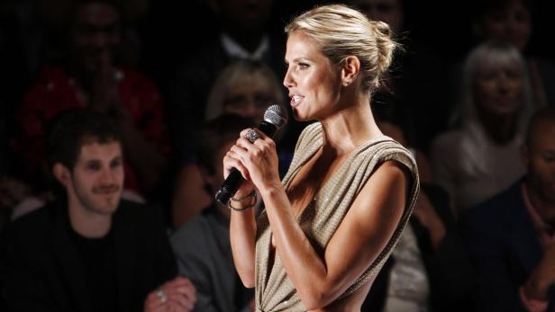 Heidi Klum spricht bei einer Veranstaltung in einem goldenen Kleid in ein Mikrofon.