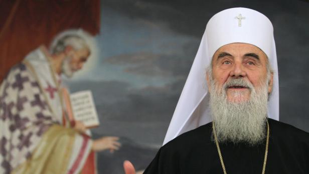 Ein orthodoxer Bischof mit weißem Bart und Mitra vor einem religiösen Gemälde.