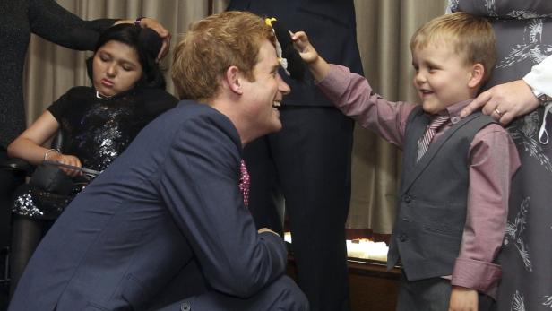 Prinz Harry kniet vor einem kleinen Jungen, der ihm ein Spielzeug ins Gesicht hält.