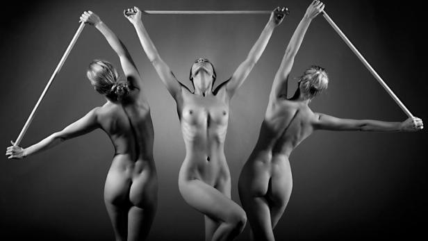 Drei nackte Frauen posieren mit einem Holzstab in einer Schwarzweißaufnahme.