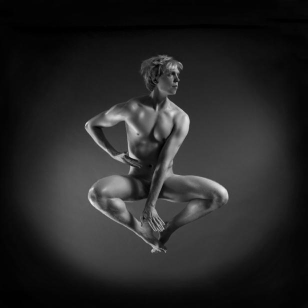 Ein junger Mann mit blonden Haaren springt in einer künstlerischen Pose vor einem dunklen Hintergrund.