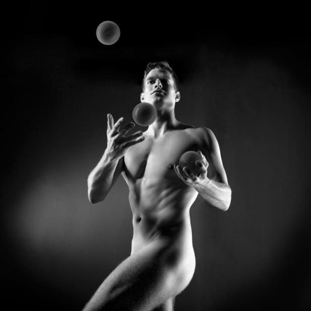 Ein nackter Mann jongliert mit drei Bällen vor einem dunklen Hintergrund.
