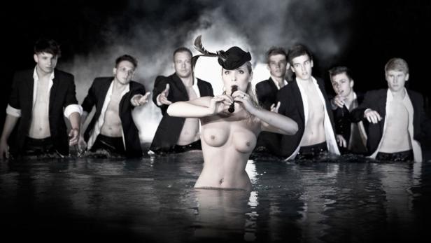 Eine Frau mit Hut spielt Flöte, umgeben von sechs Männern im Wasser.